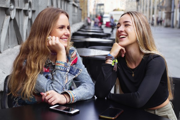Two teenage girls smile and chat outdoors, with cell phones on the table, enjoying each other's