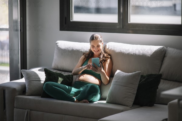 A pregnant woman sits comfortably on a cozy sofa, wearing a yoga outfit. She enjoys her leisure