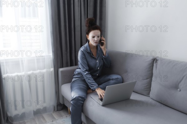 A woman in pajamas and a facial mask sits on a gray couch, multitasking between a laptop and a
