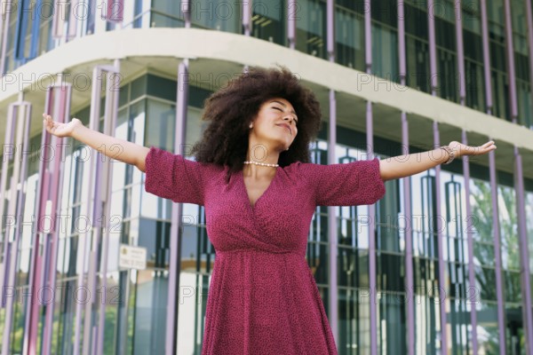 Young Arab woman with afro hair, wearing a pink dress, joyfully extends her arms in front of a