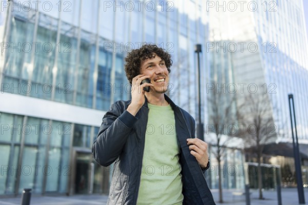 A businessman stands outside a sleek, modern glass building, smiling as he talks on his smartphone,