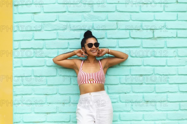 A young Latin woman stands against a vibrant turquoise wall in Mancora, Peru. Wearing a striped top