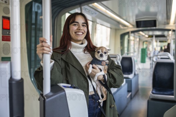 A woman smiles while holding her small chihuahua on a tram. The chihuahua looks calmly at the