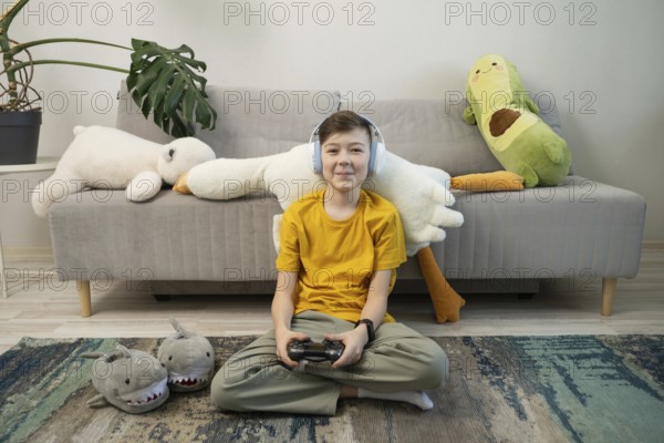 A cheerful boy in a yellow shirt and headphones plays video games, seated in a cozy room with plush