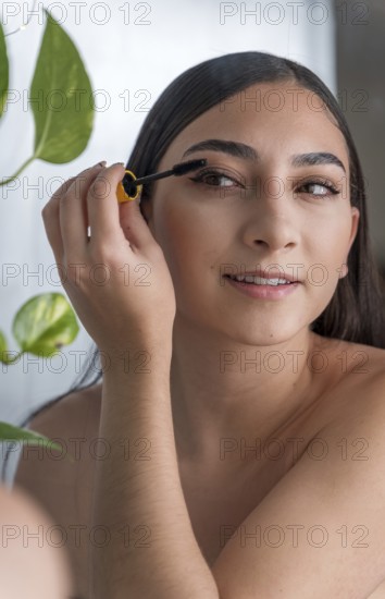 A woman smiling as she applies mascara in a brightly lit room. The presence of a vibrant green