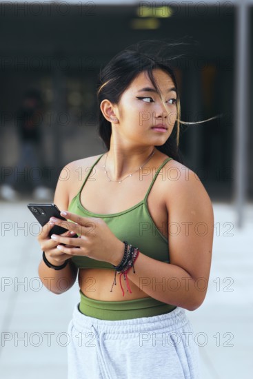 A teenage girl holds a smartphone while standing outdoors. Dressed in a green top and light pants,