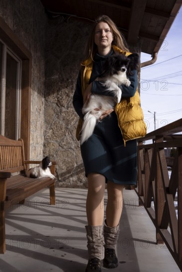 A woman stands on a rustic porch holding her purebred papillon dog. Natural light illuminates the