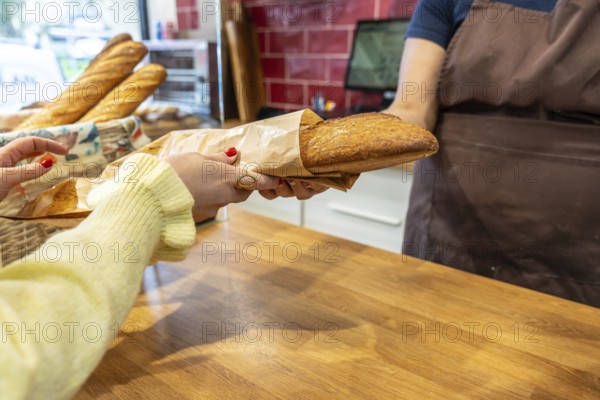Customer hands collecting a freshly baked baguette wrapped in paper from a baker behind a wooden