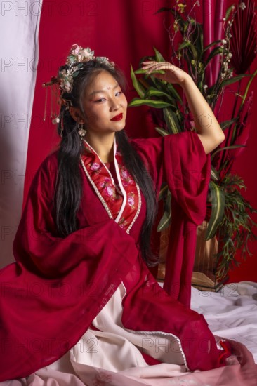 A woman in a red robe sits on a bed with a potted plant behind her. She is wearing a flower
