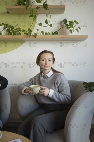 A woman sits comfortably in a cafe, sipping coffee. She is surrounded by green plants, creating a