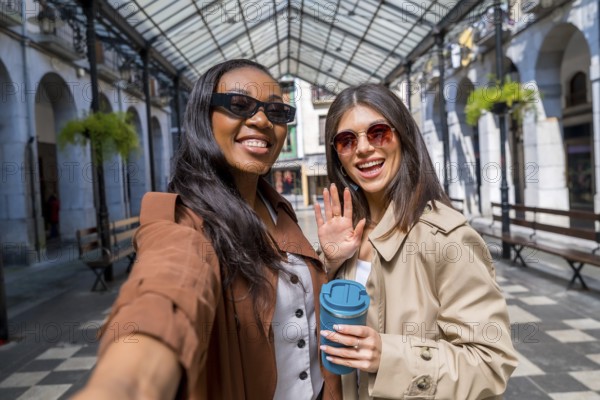 Two smiling businesswomen taking a selfie and waving at the camera in a city center gallery while
