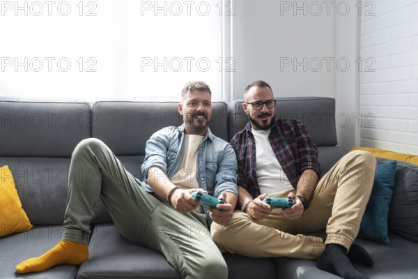 A joyful gay couple relaxes on a couch, engaging in a lively video game. Their cheerful expressions