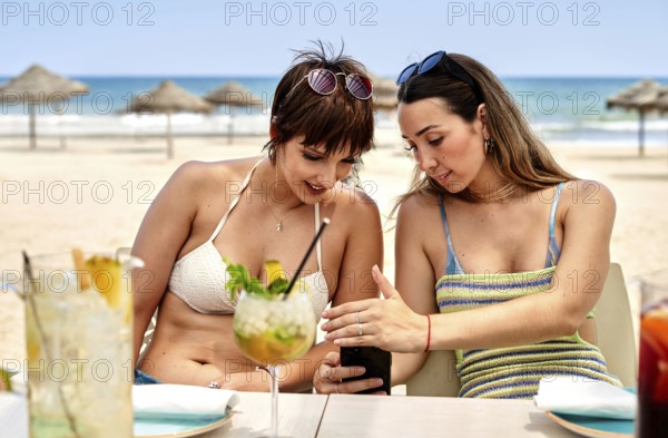 Two women sit together at a beach side table enjoying refreshing summer cocktails. Engaged in