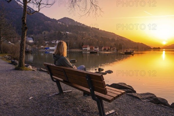 A person sitting on a bench by the lake at sunset with mountains in the background, Großer Alpsee,