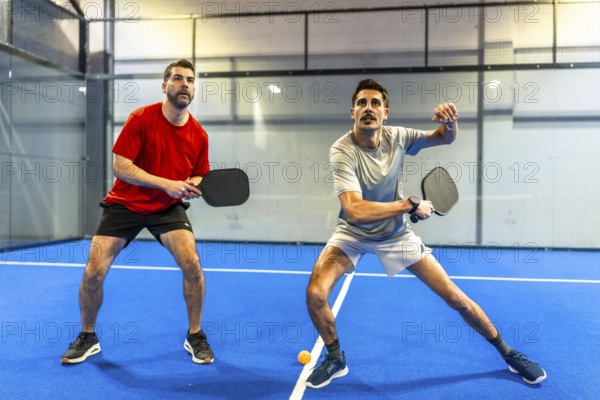 Two active men engaging in a pickleball match on a blue indoor court, concentrating on hitting the