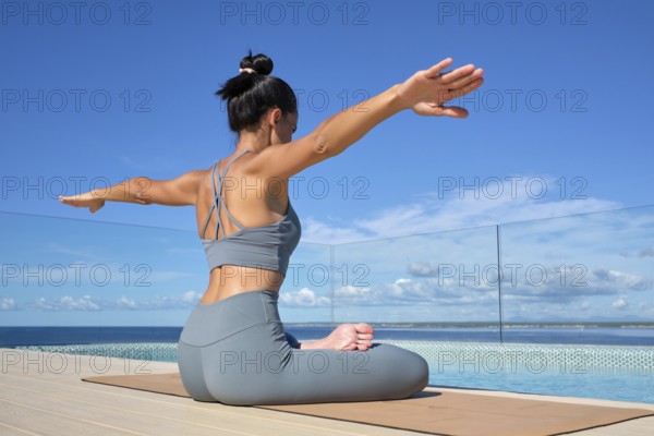 A woman practices yoga by a pool with a stunning ocean view. She sits in lotus pose on a mat,