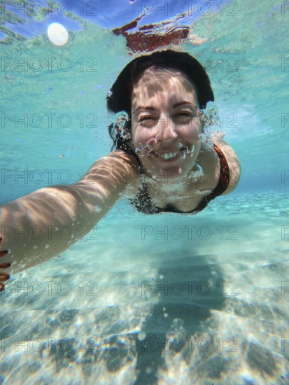 A woman swims underwater in Greece, capturing a selfie. The clear blue water reflects sunlight,