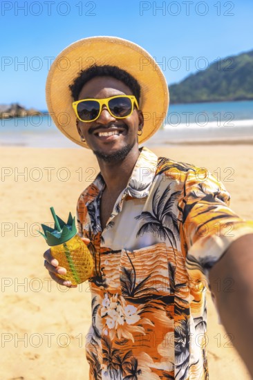 Young man taking a selfie on a tropical beach, wearing sunglasses and a straw hat, holding a