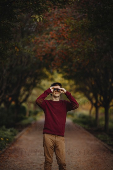 A man in a red sweater looks through binoculars amidst vibrant autumn foliage in a scenic park