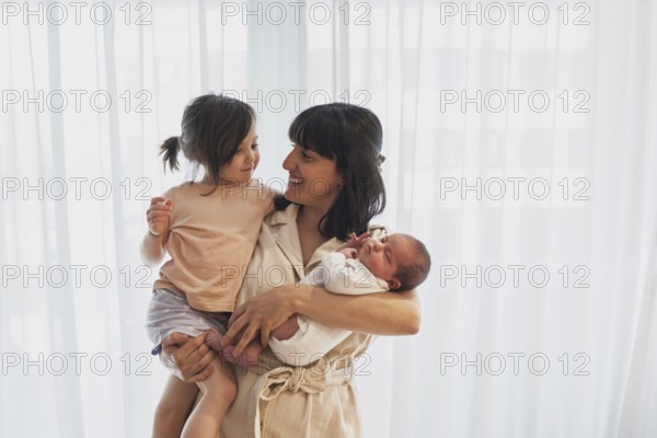A joyful young family scene with a mother holding her newborn baby and big sister. Both children