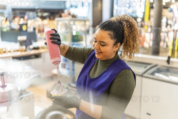Woman employee in uniform smiling as she pours pink syrup over frozen yogurt in a cup at a shop