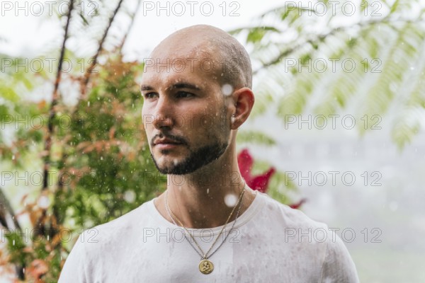 A man stands in the rain, deep in thought, surrounded by lush foliage in a forest. Drops fall