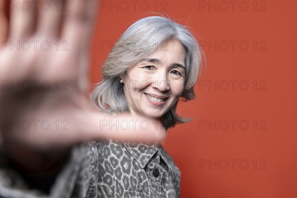 Asian senior woman smiles warmly while posing in front of a vibrant orange background. Her gray