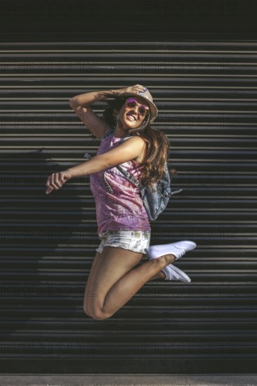 A young woman in sunglasses and a hat jumps joyfully against a dark urban background, embodying