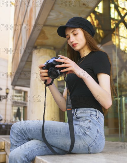 A woman photographer sits on a city bench, focused on her camera. Dressed in casual attire with a