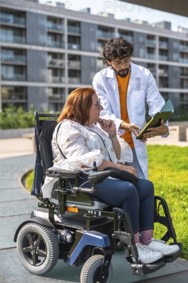 Doctor showing medical information on clipboard to woman using electric wheelchair outdoors in a