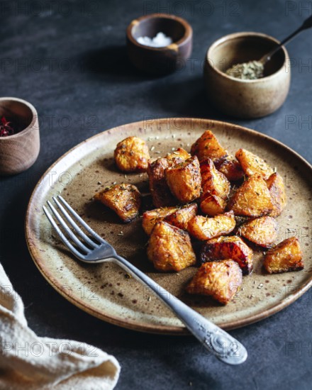 A rustic ceramic plate filled with crispy, herb roasted potatoes sits on a dark table. A fork and