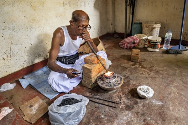 A man sits in a workshop, uses a bellows to start a fire, a man burns sapphires in a small oven in