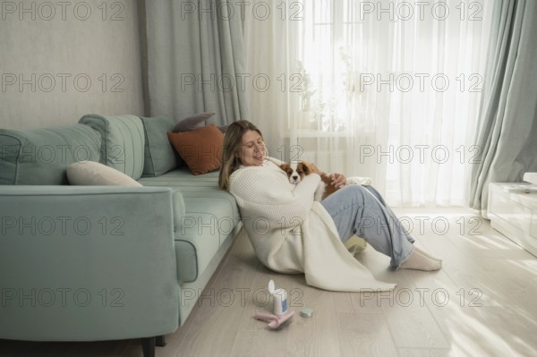 A woman cuddles her dog in a cozy home setting, sitting next to a teal sofa. Sunlight filters