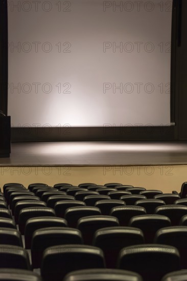 Rows of empty convention center seats facing a blank stage with a large screen, creating a quiet
