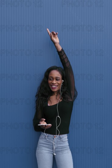 A young woman in casual attire, listening to music with earphones, raises her hand in excitement.
