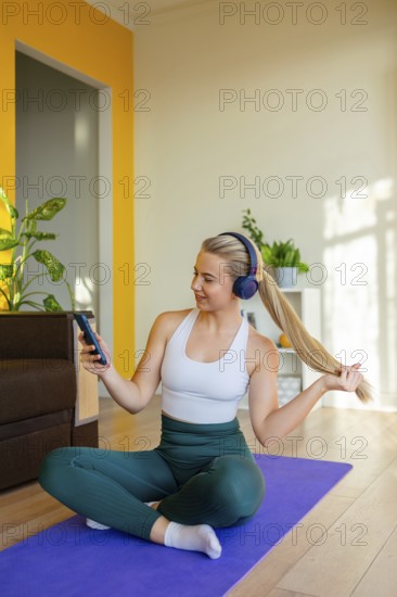 A woman in active wear sits on a yoga mat at home, wearing headphones and looking at her phone. She