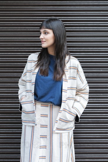 A woman stands confidently against a corrugated metal backdrop, wearing a stylish striped outfit.