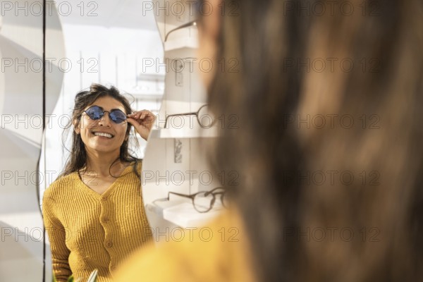 A smiling woman in a mustard sweater tries on stylish sunglasses while looking in a mirror at a