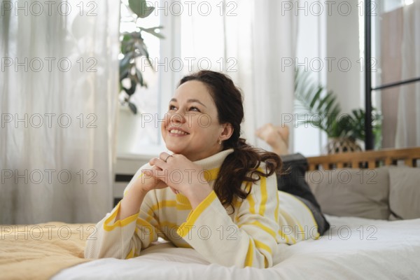 A cheerful woman in a cozy, striped sweater lies on a comfortable bed, gazing contently. The room