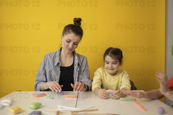 A woman and child create colorful clay shapes at a white table against a vibrant yellow background,