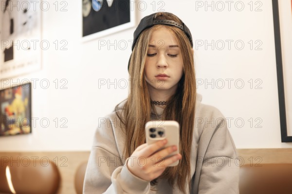 A teenage girl in a casual hoodie and cap sits indoors, focused on her smartphone. The background