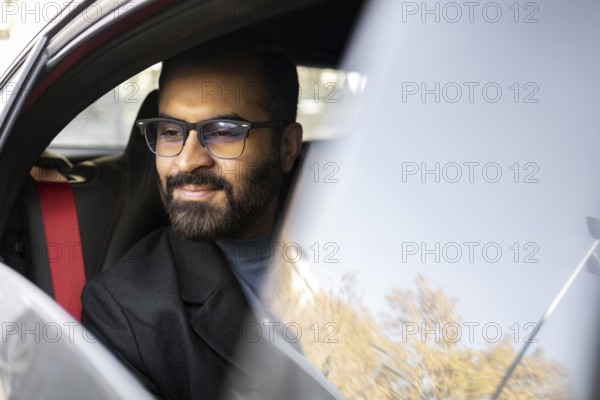 An Indian man dressed in a dark winter coat and glasses is seen smiling gently while driving his