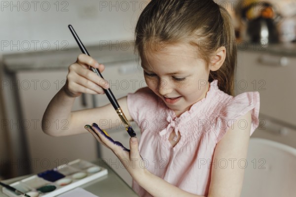 A joyful young girl in a pink top paints her hand with watercolor, showcasing creativity and fun.