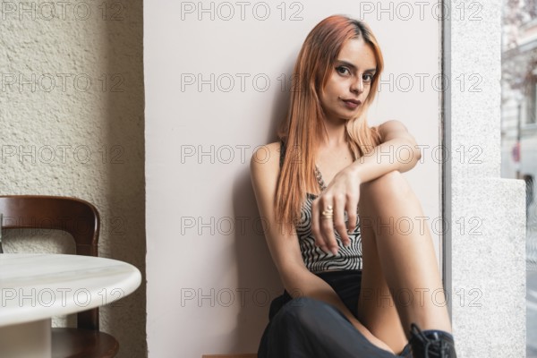 A pensive woman with orange hair sits by a window in a cafe, her expression thoughtful. Soft