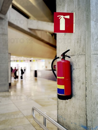 A red fire extinguisher mounted on a concrete wall in an airport terminal. A safety sign above it