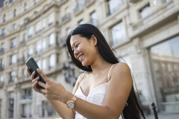 A young Chinese woman smiling while using her smartphone on a sunny summer day in the city. The