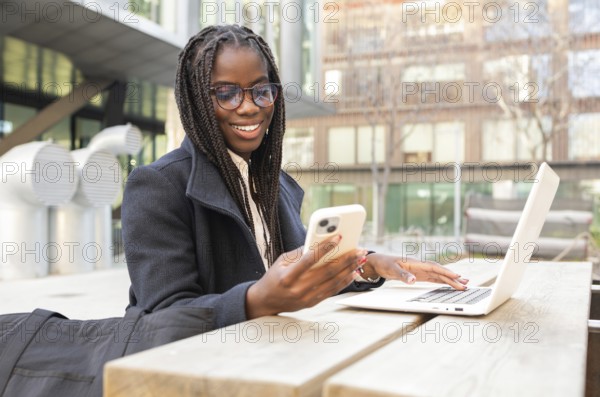 A young African American businesswoman with braids is smiling as she uses her smartphone and laptop