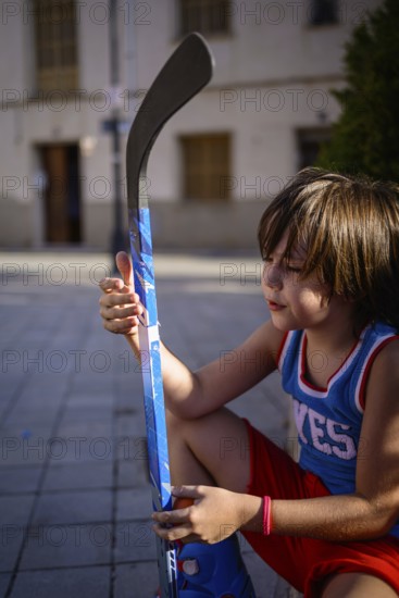 A young child sits on a paved street, holding a vibrant blue hockey stick. The scene captures a