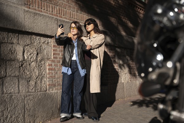 A multiethnic lesbian couple poses for a joyful outdoor selfie, exuding pride and love. The duo
