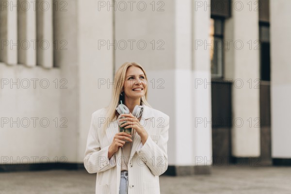 A smiling woman stands outside in an urban setting, holding a drink and headphones. She wears a